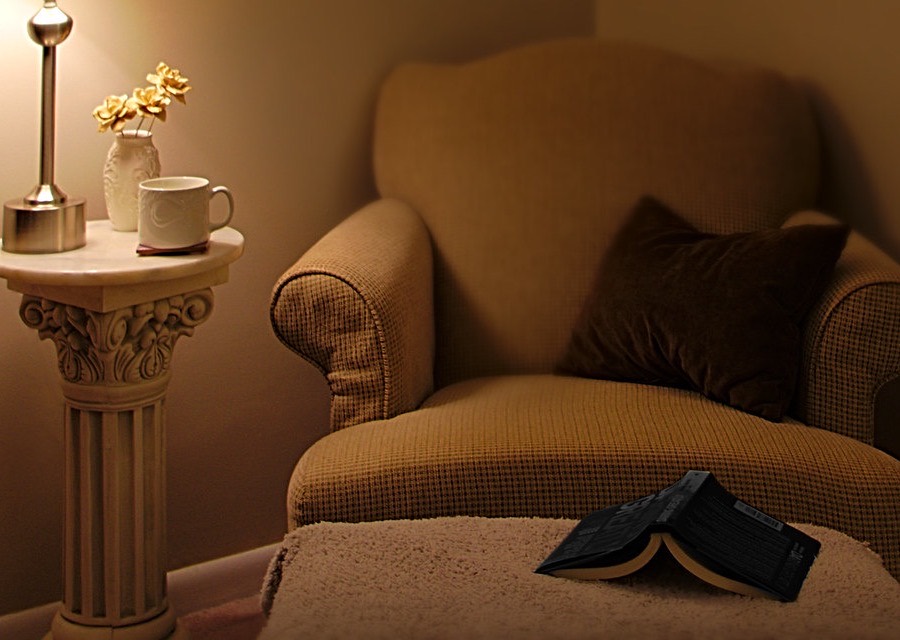 A softly lit armchair in a quiet corner, with a lamp, a cushion and an open book.